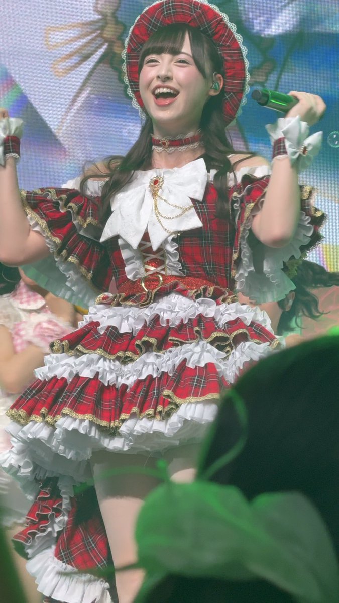 A young performer with long brown hair beams with joy while singing into a green microphone, wearing an elaborate red plaid frilly dress and matching bonnet with lace trim, consistent with the post celebrating "のあちゃん" (Noa-chan) singing "のびグリ" (Nobiguri) with care during a live performance.