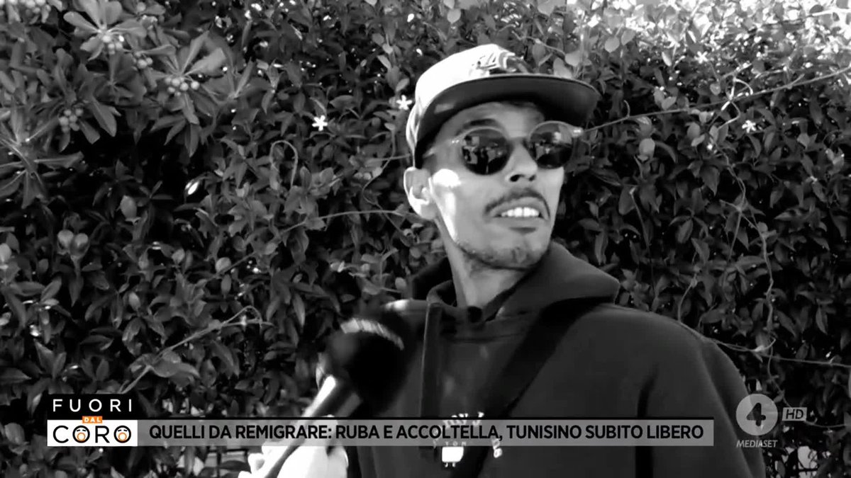 A black-and-white screenshot from Italian TV show "Fuori dal Coro" features a man wearing a cap and round sunglasses being interviewed outdoors against a leafy background, with the on-screen headline reading "QUELLI DA REMIGRARE: RUBA E ACCOLTELLA, TUNISINO SUBITO LIBERO." The accompanying post quotes the interviewed man's confession, identifying him as Jamil, the Tunisian immigrant who allegedly robbed and stabbed someone in the Marche region before being released, a case the show is using to argue for stricter deportation policies.