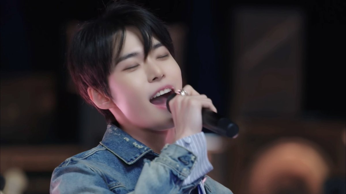 A young man with dark hair sings passionately into a microphone while wearing a denim jacket.