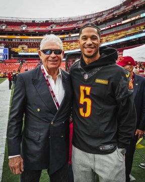 Two men pose together on the football field at a stadium, with one wearing a black Washington Commanders jersey featuring the number 5 and the other dressed in a dark blazer and sunglasses. The image relates to the post by showcasing the trio mentioned—Josh Hart in his Commanders #5 jersey alongside the legendary "Riggo" (former Redskins running back John Riggins).