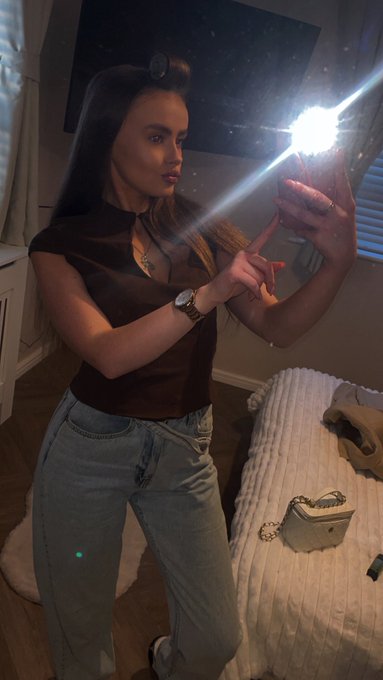 A woman takes a mirror selfie in a bedroom using a phone with the flashlight on, wearing a brown top and blue jeans.