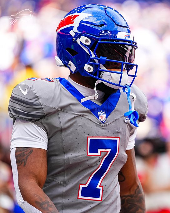 A Buffalo Bills football player wearing jersey number 7 and a blue helmet stands on the field during the Sunday afternoon matchup against the Los Angeles Chargers. His helmet features the Bills logo, and his gray jersey with blue accents displays the NFL shield, connecting to the #LACvsBUF game day setting.