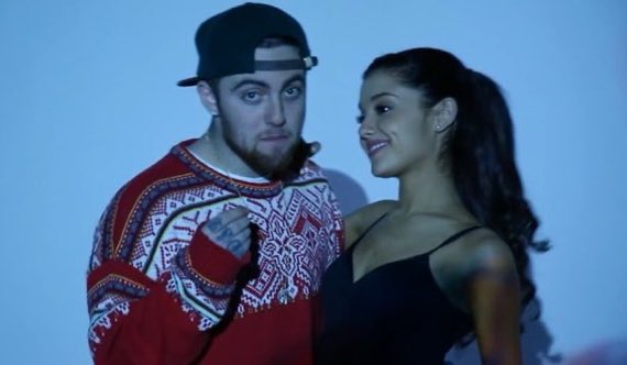 A man in a backwards cap and patterned sweater points at himself while standing next to a smiling woman with long dark hair in a black top, against a blue-lit background. The image accompanies the news that Ariana Grande and Mac Miller have songs simultaneously charting in the Top 10 on the US Rhythmic Radio chart, recalling their 2013 collaboration "The Way."