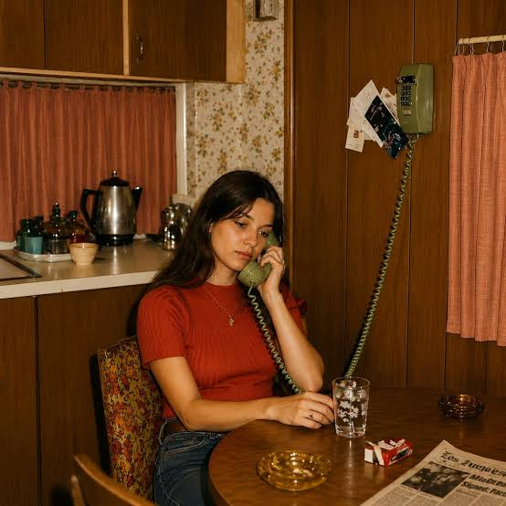 A young woman in a red knit top talks on a green rotary wall phone at a wood-paneled kitchen table featuring a "Los Angeles" newspaper. The scene is intentionally styled to look like a 1970s photograph, though subtle modern anachronisms would be hidden in plain sight to test the viewer's eye.