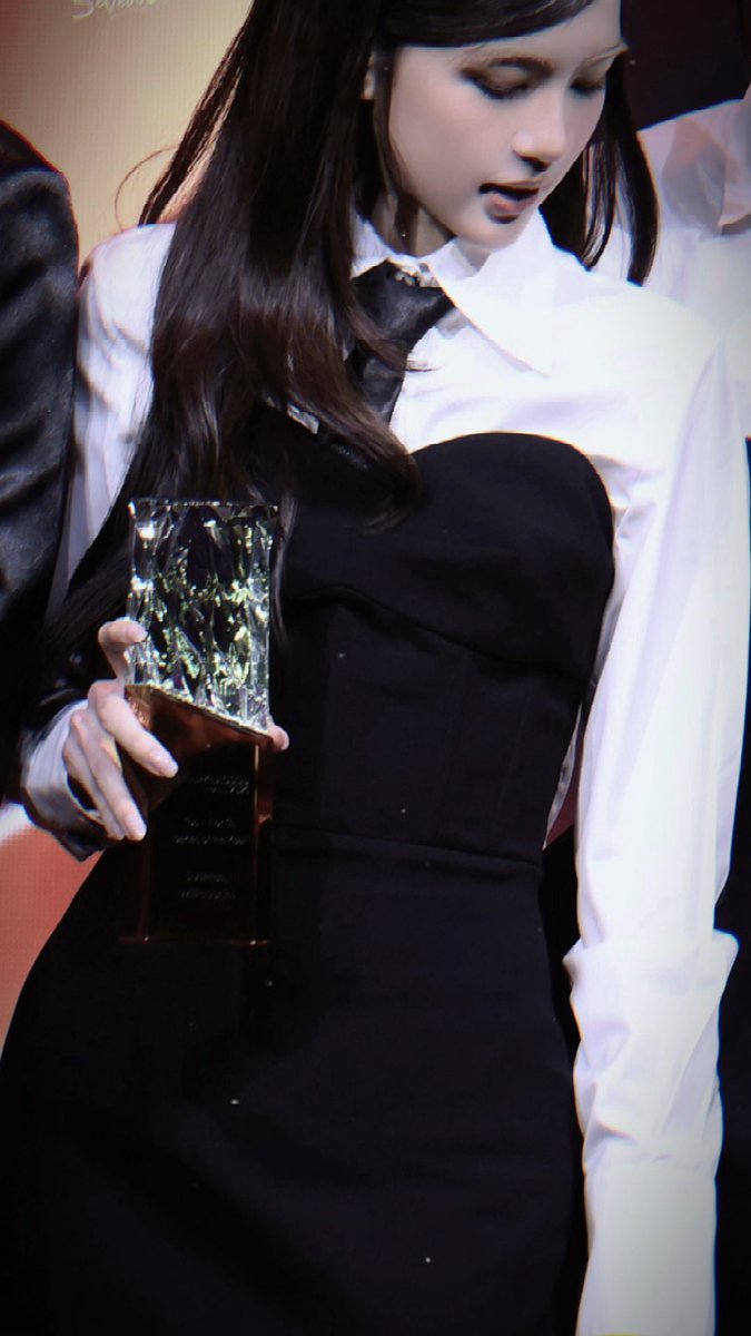 A young woman with long dark hair wearing a white blouse and black dress, looking down at a crystal trophy she is holding.