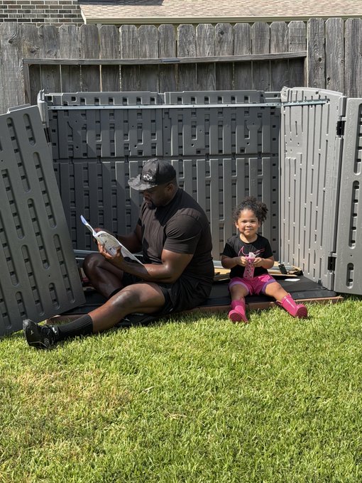 An adult man in a black cap and t-shirt reads instructions while sitting on the partially-assembled gray panels of a shed in a backyard, with his smiling young daughter in pink boots sitting beside him "helping" with the project. The image perfectly captures the heartwarming dad-and-daughter teamwork moment described in the post about putting a shed together.