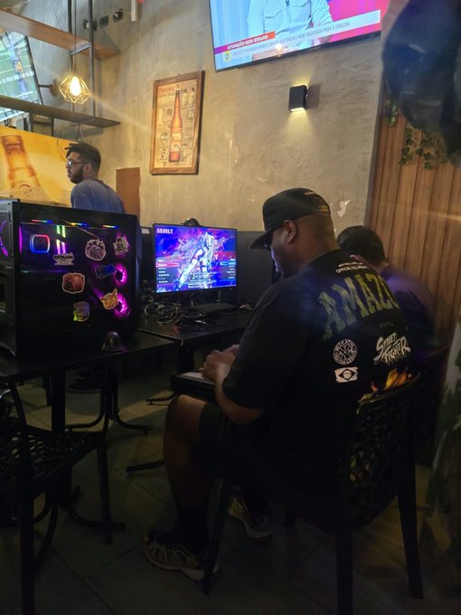 A person wearing a black 'Street Fighter' cap sits at a desk playing a video game on a computer with RGB lighting in a cybercafe setting.
