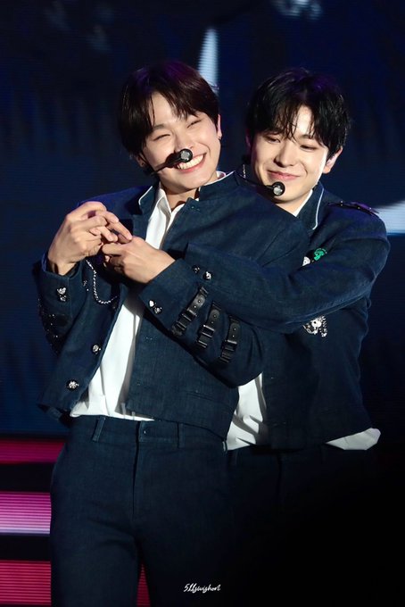 Two male performers wearing matching dark denim outfits with headset microphones share a playful, close embrace on stage, one smiling broadly while the other winks with a scrunched expression, perfectly capturing the cute and affectionate "Kuukuuz face mashup" energy described in the post.