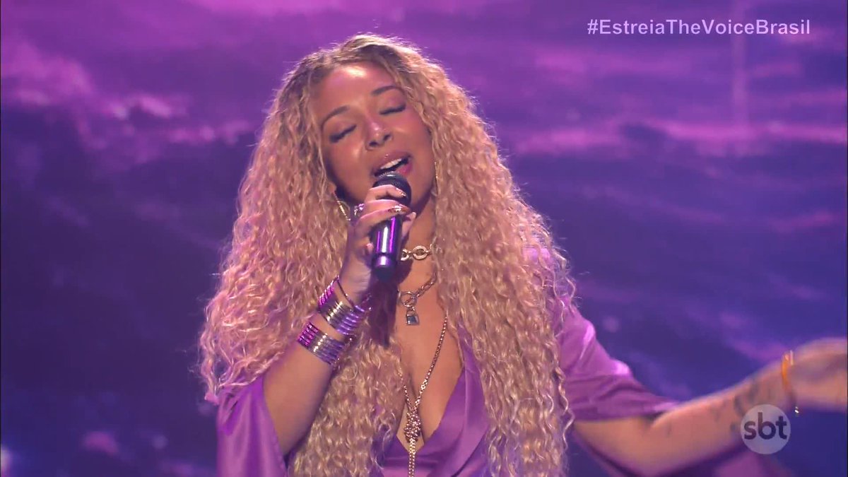 A blonde woman with long curly hair sings passionately into a microphone against a purple-hued background, wearing a silky purple blouse and accessorized with a padlock pendant necklace and multiple bracelets. The hashtag "#EstreiaTheVoiceBrasil" appears in the upper right corner, and the "sbt" logo is visible in the bottom right, fitting the post about Lia Lira's premiere performance of "Purple Rain" on The Voice Brasil.