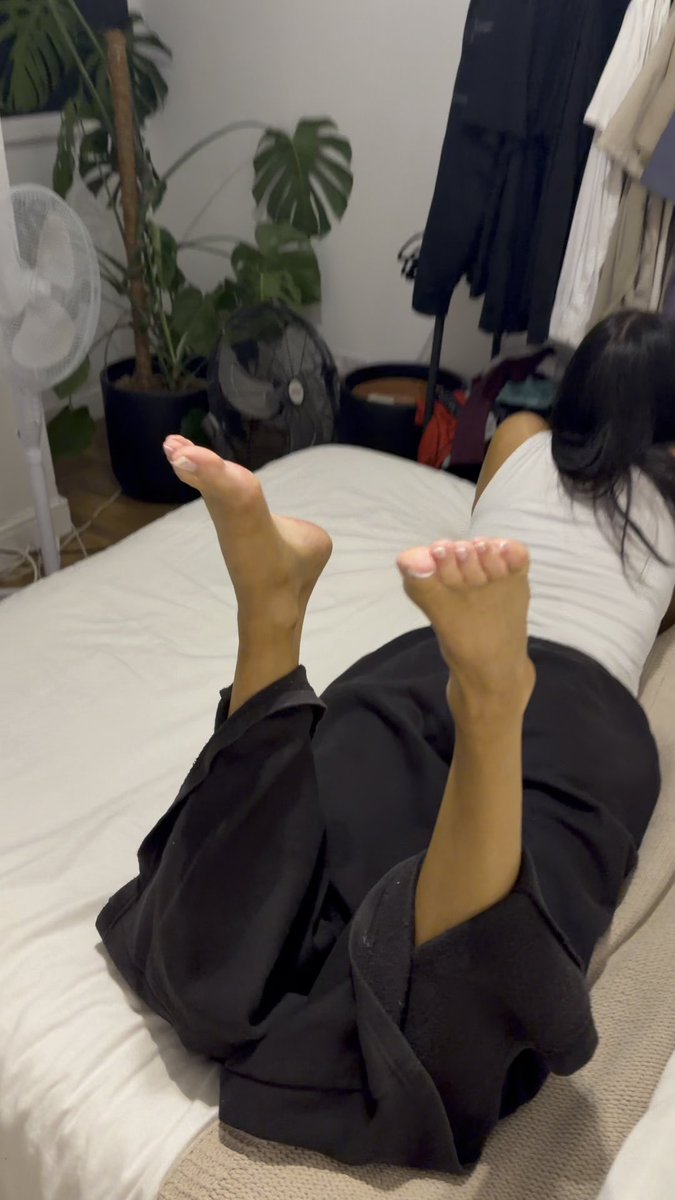 A photograph taken from behind of a person lying face down on a bed, wearing a white top and dark pants, with their bare feet raised in the air.