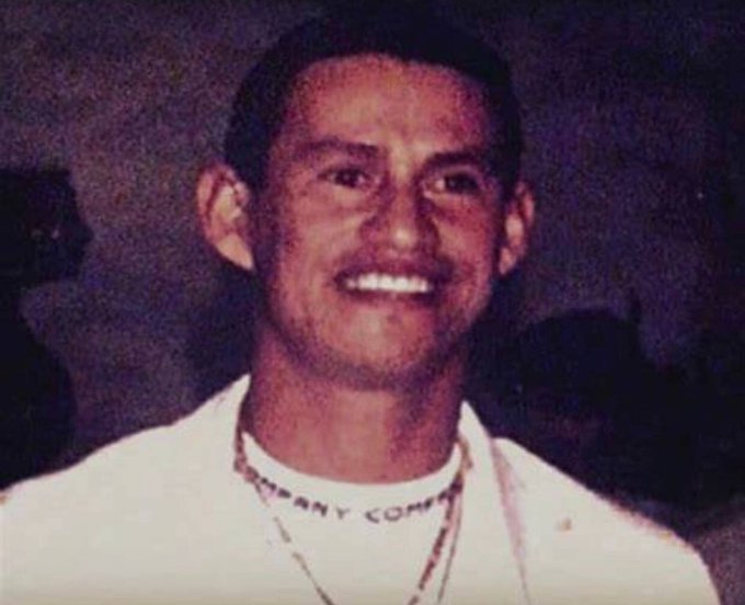 An old photograph of a smiling man with a mustache wearing a white collared shirt with a necklace, against a dark background.