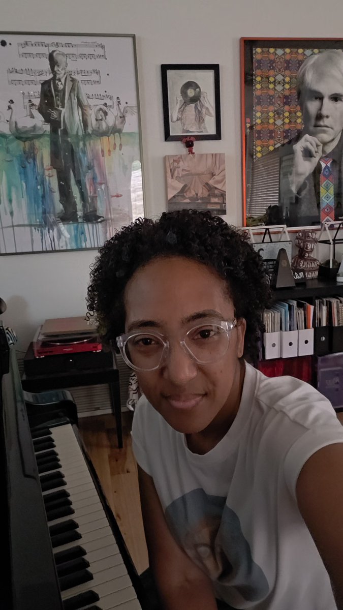 A person with curly hair and clear glasses takes a selfie in a room decorated with a piano, bookshelves, and various framed artworks, including a portrait resembling Steve Jobs.