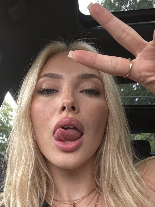A blonde woman taking a selfie in a car, making a peace sign and sticking out her tongue, wearing a gold ring and a necklace.