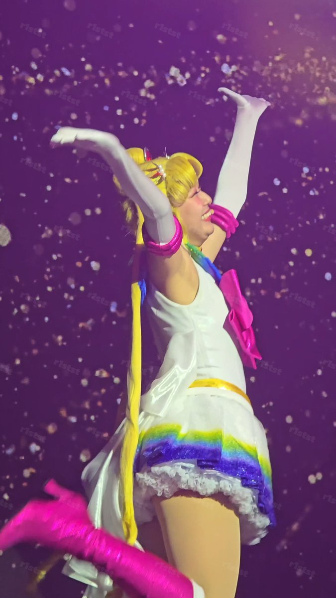 A performer in a Sailor Moon costume with blonde pigtails, white gloves, and pink accents raises her arms joyfully against a sparkling purple background.