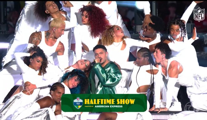 An image promoting the 'HALFTIME SHOW PRESENTED BY AMERICAN EXPRESS' shows a performer in a green outfit surrounded by dancers in white at an NFL event.