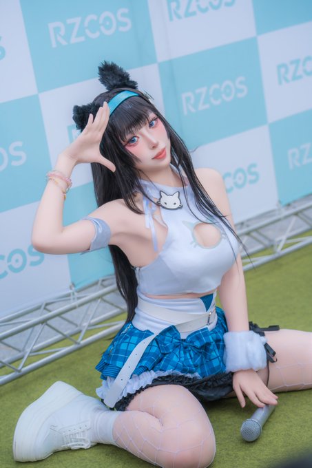 A cosplayer dressed in a blue and white outfit with cat ear accessories poses at an event with an 'RZCOS' banner backdrop.