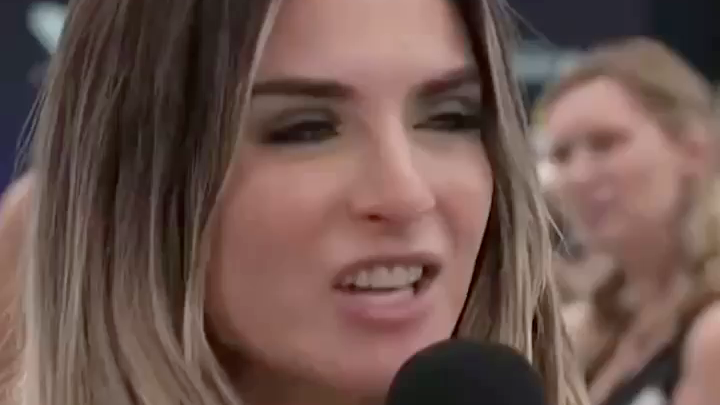 Close-up of a woman with long brown hair speaking into a microphone, appearing to be at a public event or interview.
