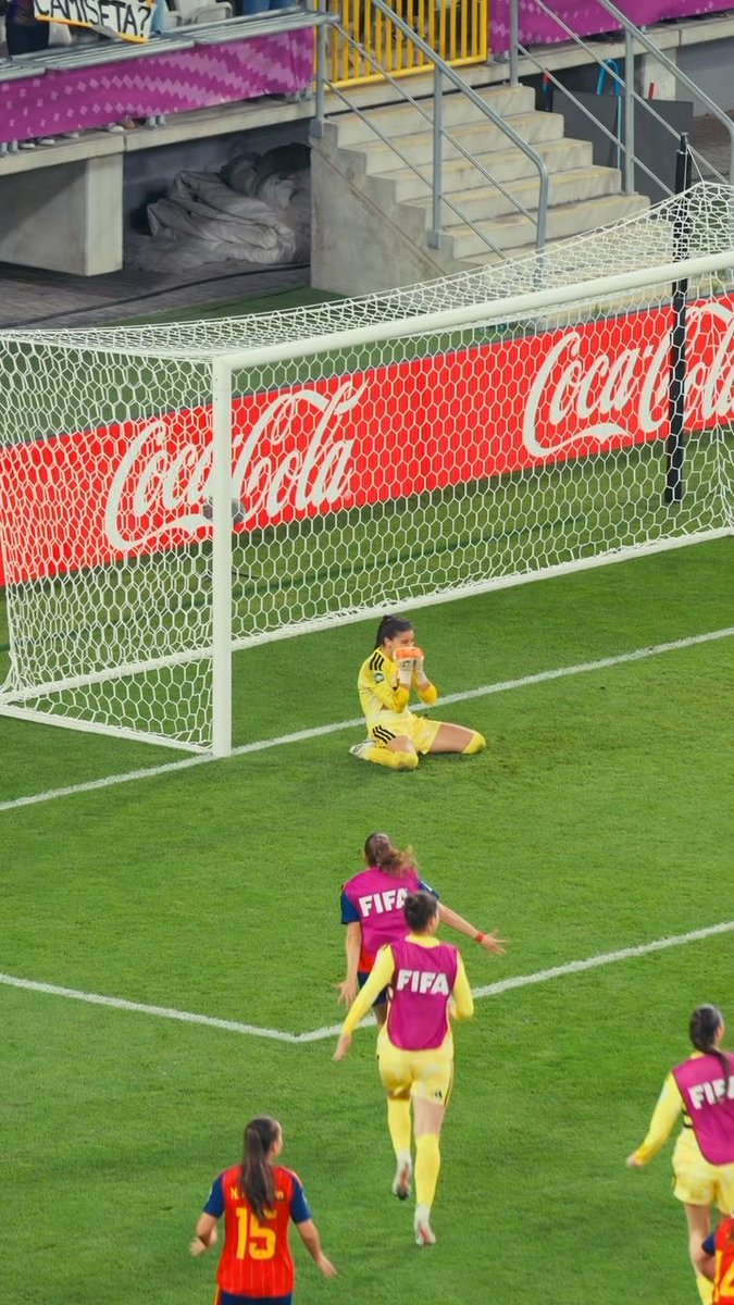 A goalkeeper in a yellow kit sits on the ground in front of the goal net, appearing to celebrate, while several players wearing pink "FIFA" bibs run toward her on the pitch. The scene captures the key moment referenced in the post—the decisive save by Laia López that secured Spain's championship at the #U20WWC, with Coca-Cola advertising boards visible behind the goal.