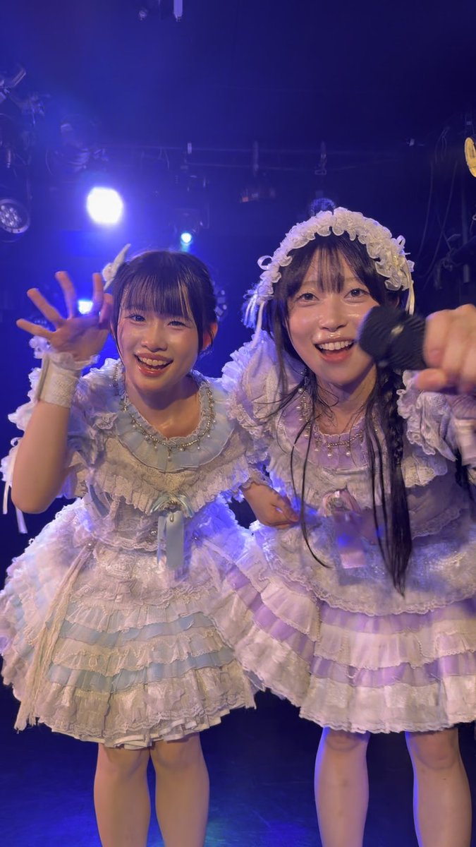 Two performers in elaborate lavender and white ruffled costumes smile warmly at the camera in a dimly lit venue with blue stage lighting.
