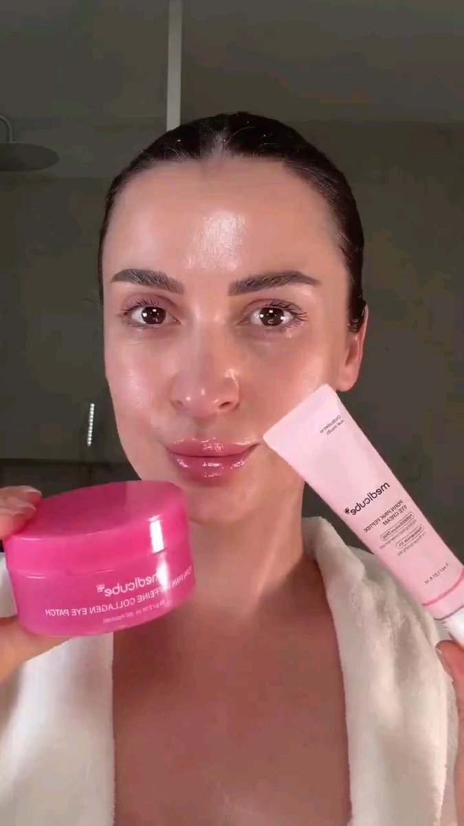 A woman with slicked-back hair and glossy skin holds up two Sol de Janeiro beauty products, a pink eye batch pot and a Brazilian Bum Bum cream tube.