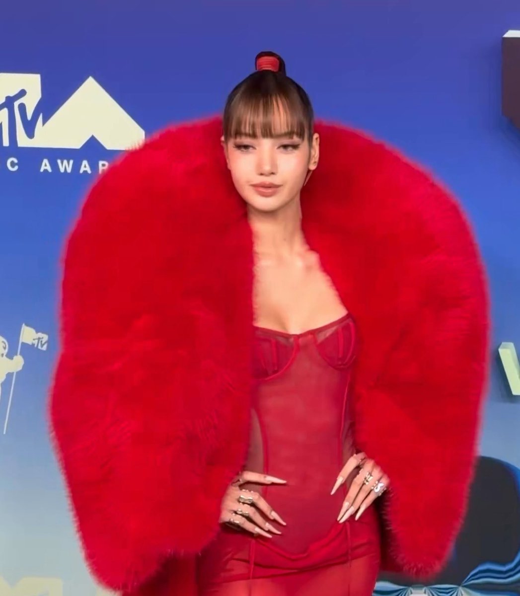 The young woman from the first image poses on the MTV Awards blue carpet in her red fur coat and sheer red corset dress, showing her long nails and rings.