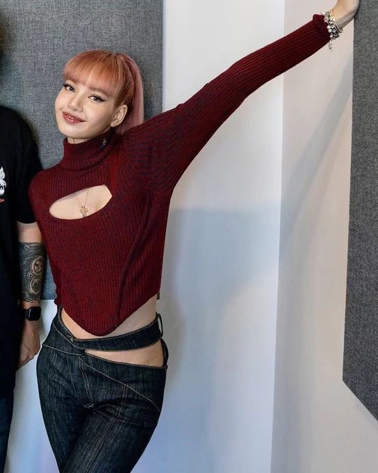 A smiling woman with pink hair wears a maroon cutout turtleneck and dark jeans, posing with one arm raised.