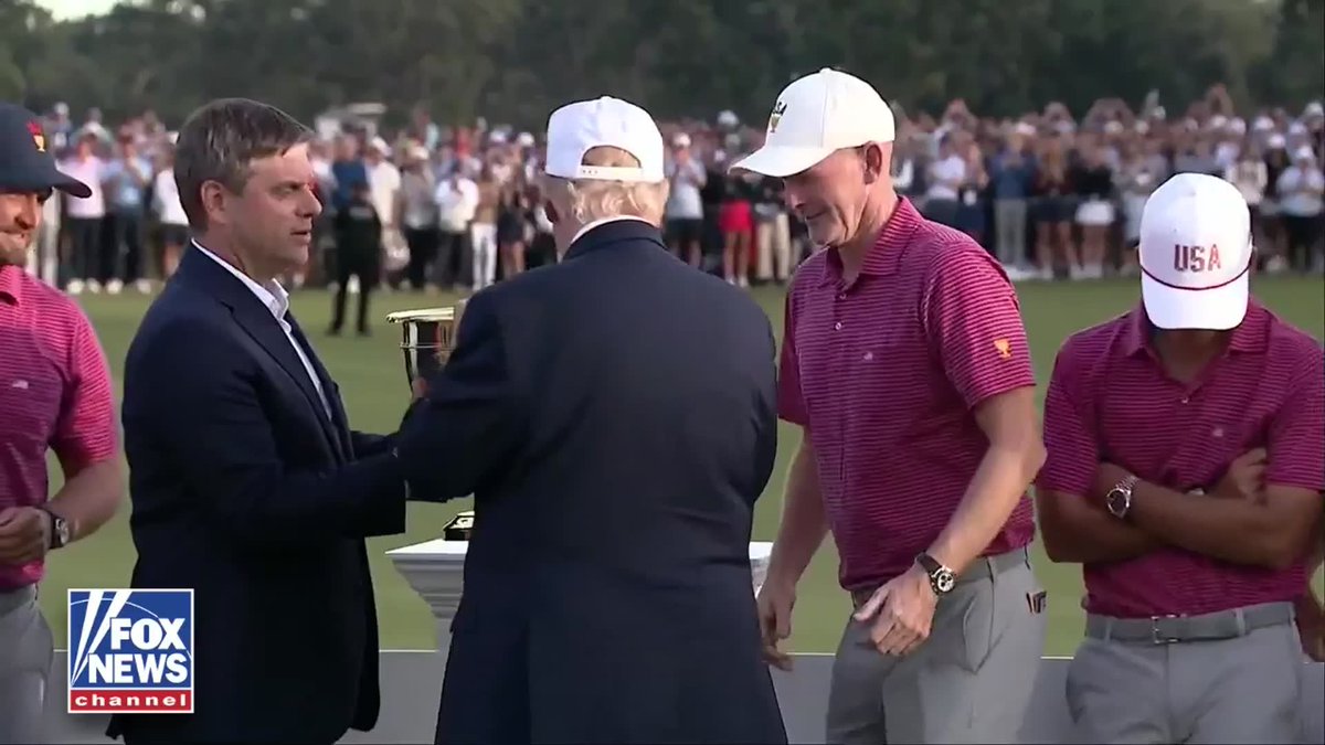 President Trump, wearing a "USA" cap, hands the Presidents Cup trophy to U.S. Team Captain Brandt Snedeker as American golfers in matching maroon polos celebrate their 17-13 victory over the International Team at Medinah Country Club, with the "Fox News Channel" logo visible in the corner.