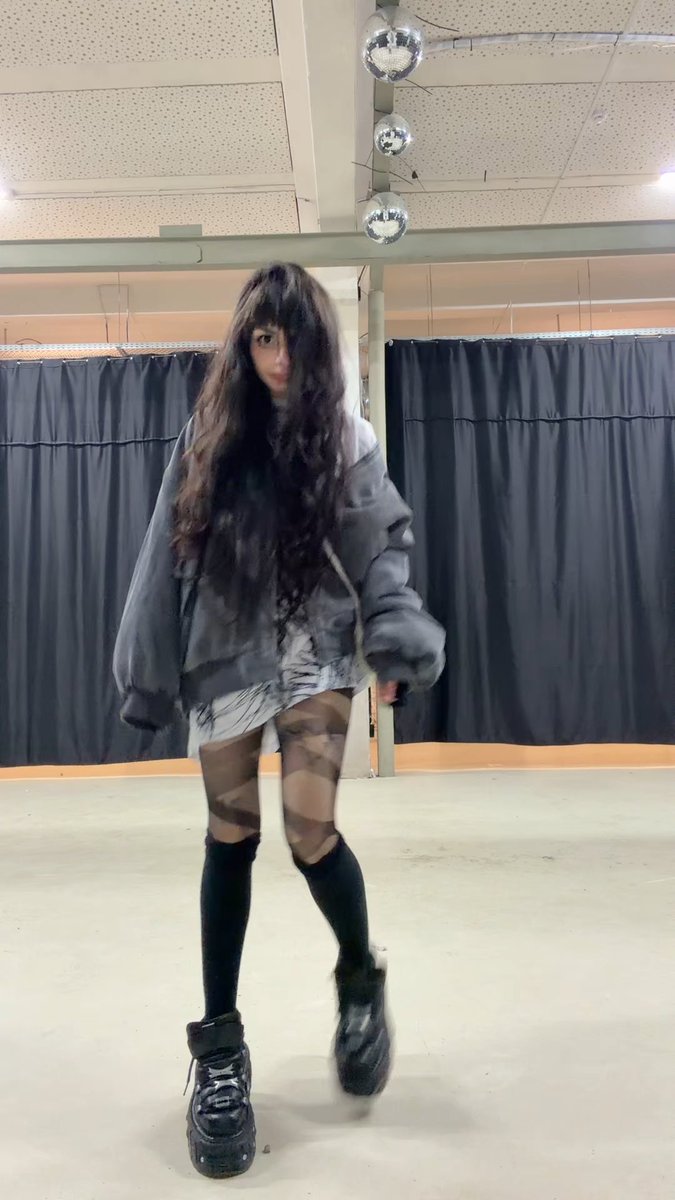A person with long dark hair covering their face, wearing an oversized gray jacket, tights, knee-high socks, and black shoes, posing in a dance studio with disco balls.