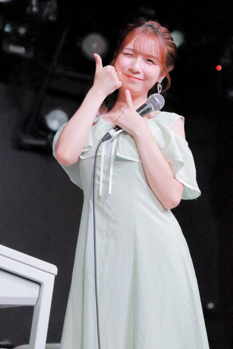 A young woman with light brown hair, wearing a pale green dress, holds a microphone and gives a double thumbs-up gesture on stage.