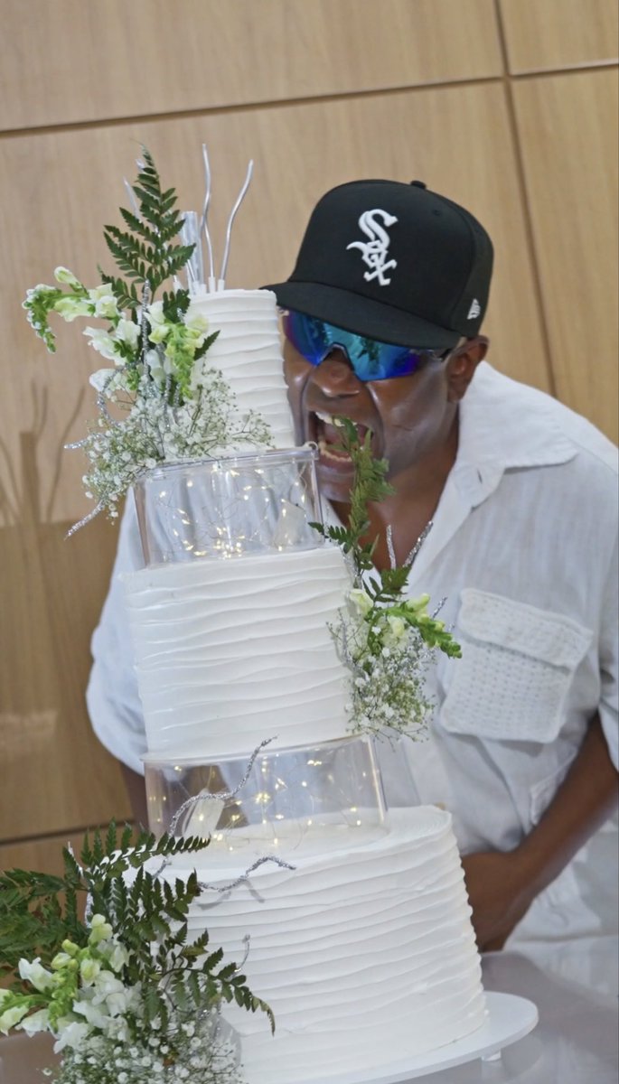 A man wearing a Chicago White Sox cap and blue reflective sunglasses bites into the greenery of a tiered white wedding cake decorated with ferns and baby's breath.