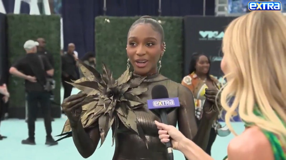 Normani is being interviewed by ExtraTV on the MTV VMAs red carpet, holding an Extra microphone while wearing a striking sheer black outfit with a dramatic leaf-like floral embellishment. The "Extra" logo is visible in the top right corner and on the microphone, while "VMA" signage appears in the background.