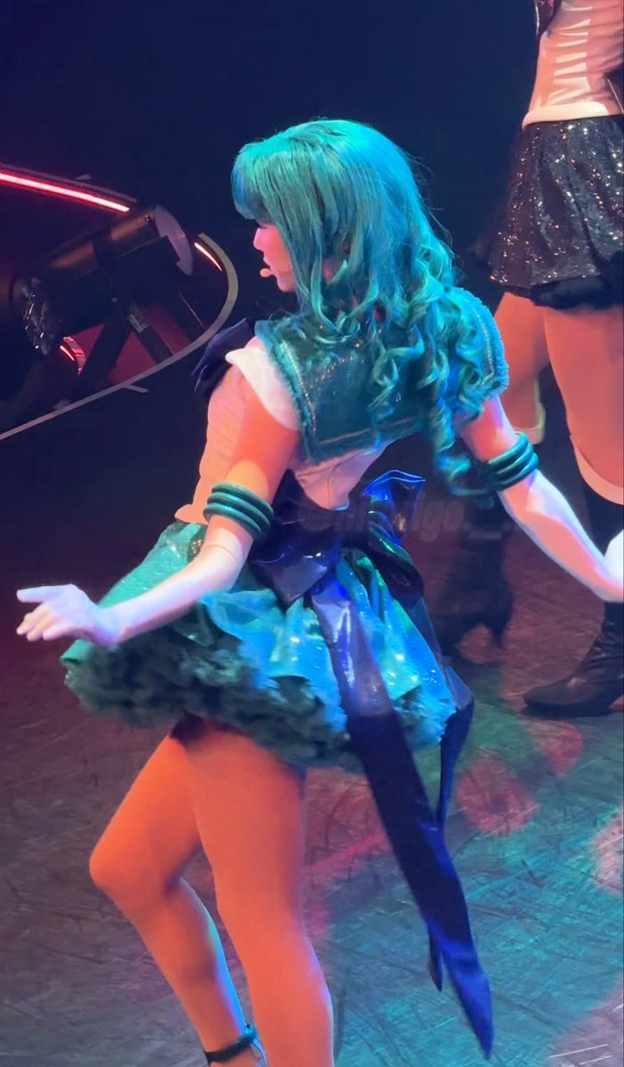 A performer with teal hair in a matching teal and white costume performs on stage, with a 'jalgo.' watermark visible.