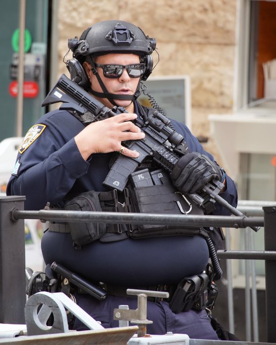 A heavily equipped NYPD Counterterrorism Bureau officer stands guard with an HK416 rifle in hand, positioned behind a metal barricade to secure the perimeter of the UN General Assembly. The setup reflects heightened security measures in New York City, with no visible text or identifying numbers on the officer's gear beyond a small "7C" marking on a pouch.