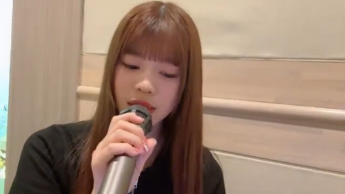 A young woman with long brown hair and bangs, wearing a black top, holding a silver microphone to her mouth with her eyes closed.