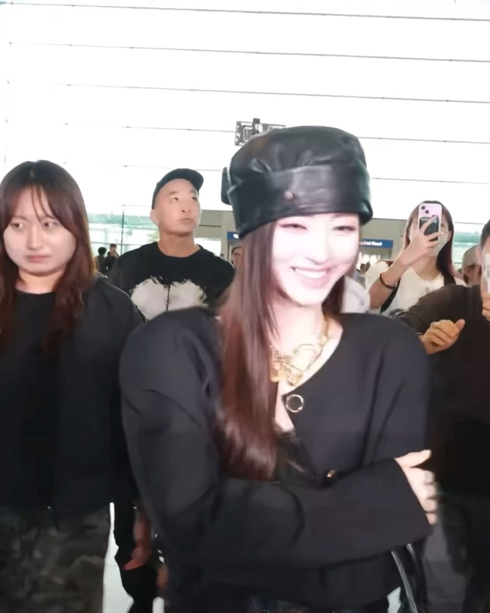 A woman wearing a large black leather hat and a black top with a gold necklace smiles while walking through what appears to be an airport, surrounded by other people including someone taking a photo with a phone.
