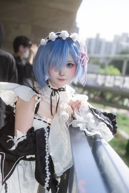 A cosplayer with short blue hair and a floral headband poses in a black and white frilly dress, leaning against a railing outdoors.