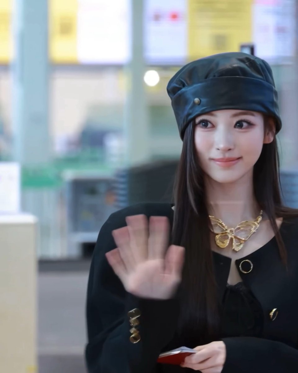 A young woman wearing a black beret and black outfit with a gold bow necklace, waving at the camera in what appears to be an airport or transit area.