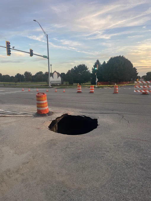 A large, deep sinkhole in the asphalt roadway at the intersection of 95th Street and Book Road. An orange and white striped traffic drum sits adjacent to the edge of the hole. In the background, traffic signals, warning cones, and detour barricades block off lanes beneath an early evening sky.