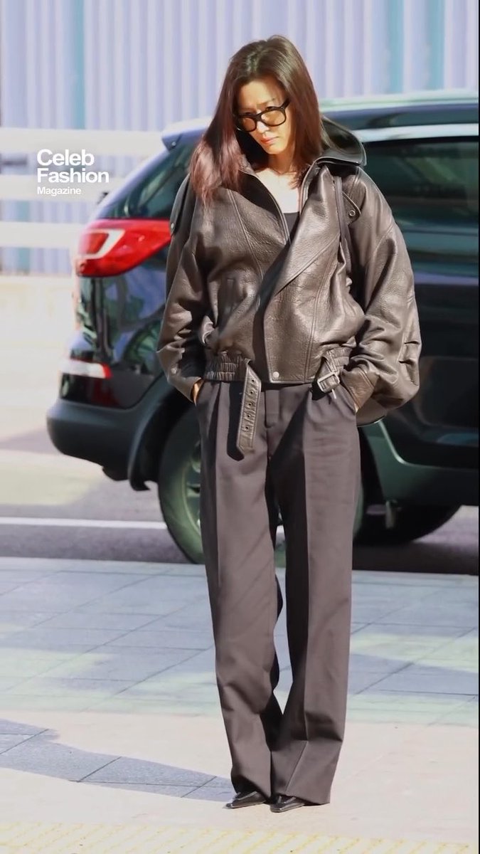 A woman in a brown leather jacket, black trousers, and glasses stands on a sidewalk with a dark SUV parked behind her, labeled 'Celeb Fashion Magazine.'
