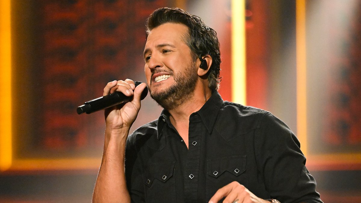 A bearded country singer, resembling Luke Bryan, smiles while singing into a microphone, wearing a black western shirt on a warmly lit stage.