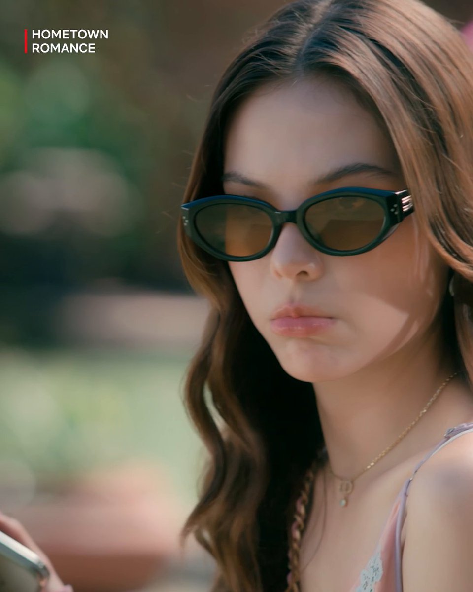 A close-up portrait of a woman with long brown hair wearing dark oval sunglasses, with the text "HOMETOWN ROMANCE" displayed in the upper left corner.