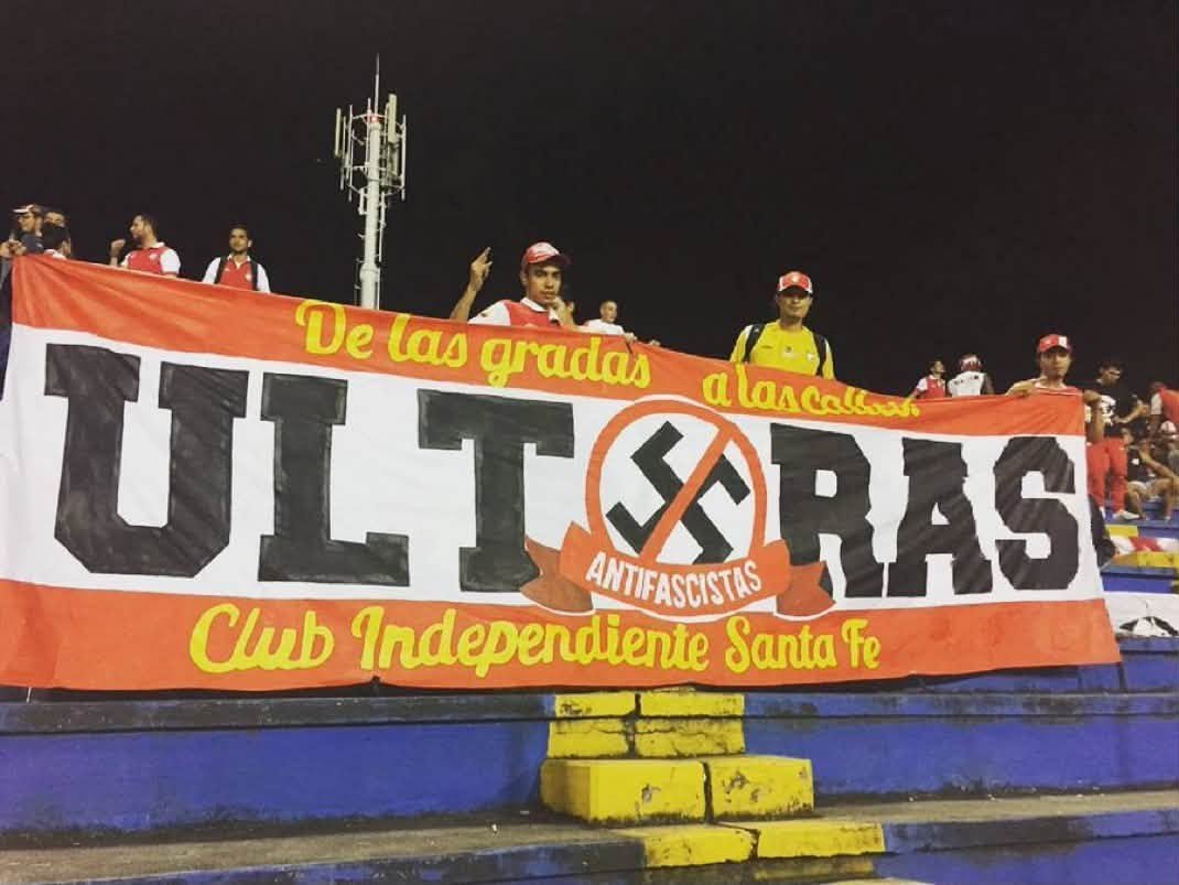 ultras_antifaa's tweet photo. Red Guards United is the anti-fascist supporters’ group of Independiente Santa Fe (Colombia).

The story of the group, in their own words:

We are Red Guards United, fervent fans of the glorious Independiente Santa Fe. Starting from counter-cultures and thus from militant anti-fascism, we consider a convergent way of endurance and carnival, with our own ideologies of the youth who does not believe in the bullshit of the media and assumes his life critically and constantly, growing personally and as a group.

We hold our fists up, with the desire to encourage and cheer and give everything of ourselves. We want to win our rights, stop being spectators and become actors in our own lives. Therefore, radicalism is also present in each of our actions, both inside and outside the stadium.
We identify ourselves as a social gesture that resists the oligarchy, the current economic system, the values of capitalism and modern football, which aims to fill the pockets of a few, leaving passion aside, and therefore, our team: Santa Fe.

In the reality in which we live, we have come to find in the streets and the stands, as anti-fascist fans, a passion with a common history, the construction of a dignified life, with a true character of justice in peace and, of course, the unity of Latin American peoples.

In this space, we think of a different way of seeing and living football, without being limited to merely observing it in a “sportive” manner, but understanding it as a scene where passion, reason and action are not separated, where what motivates us is the love for Santa Fe.

Whether our team is at home or away, in good times and bad, we do what we do best: dress our historic grandstand in red and white at every match and make it a party, a party that the fascists will never enjoy.
Added to this responsibility with Bogotá as young people, with the country in every neighborhood, every village, in every ghetto, juvenile expression of meaning through art, music, hooliganism, theater, football, politics, anti-fascism and, if necessary, at the barricade.

Finally, regarding them as an indispensable part of our history, we want to acknowledge the efforts of individuals and organizations who daily resist the murderous monotony of this dictatorship hidden under a cloak of false democracy, assuring that we will continue the work the people have entrusted to us and the responsibility that we assume every day with more commitment and resistance.