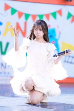 A young woman in a white dress kneels on a stage, holding a microphone and making a peace sign with her hand.