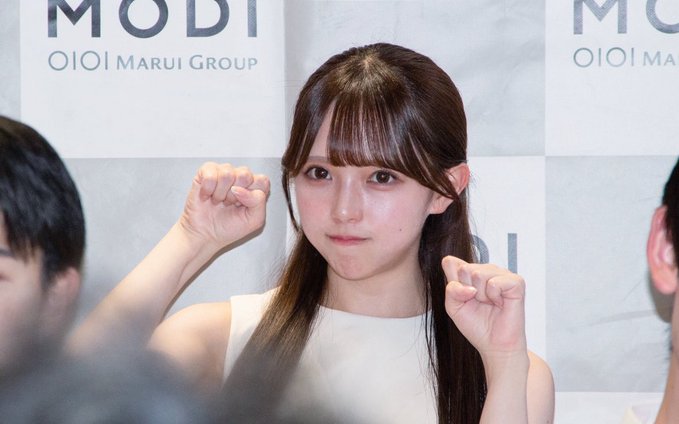 A young woman with long brown hair poses with both fists raised at an event, with a backdrop displaying 'MODI OIOI MARUI GROUP'.