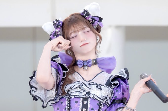 A young performer in an ornate purple and white frilly dress with matching cat-ear hair bows poses with her eyes closed and fists raised near her face while holding a microphone, appearing to be at the fav me Lily Event at Lalaport Toyosu on September 2, 2026. The image captures AbeKaren in an adorable, dreamy moment consistent with the fan event's idol-style atmosphere.
