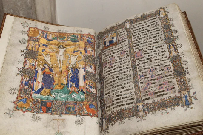 A large, ancient bound book open at a double page with an intricate illustration of the crucifixion on the left-hand side, and a page of text with an illuminated boarder on the right