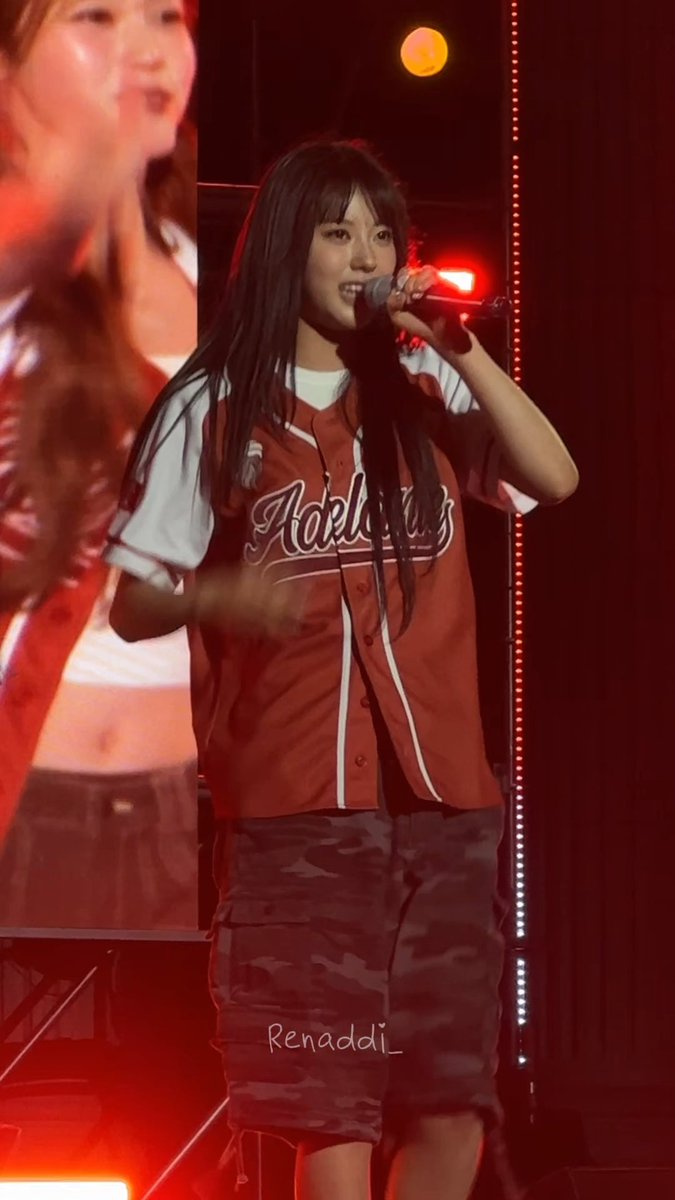 A young woman with long dark hair performs on stage wearing a red Adidas baseball jersey and camouflage cargo skirt, holding a microphone with red stage lights in the background.