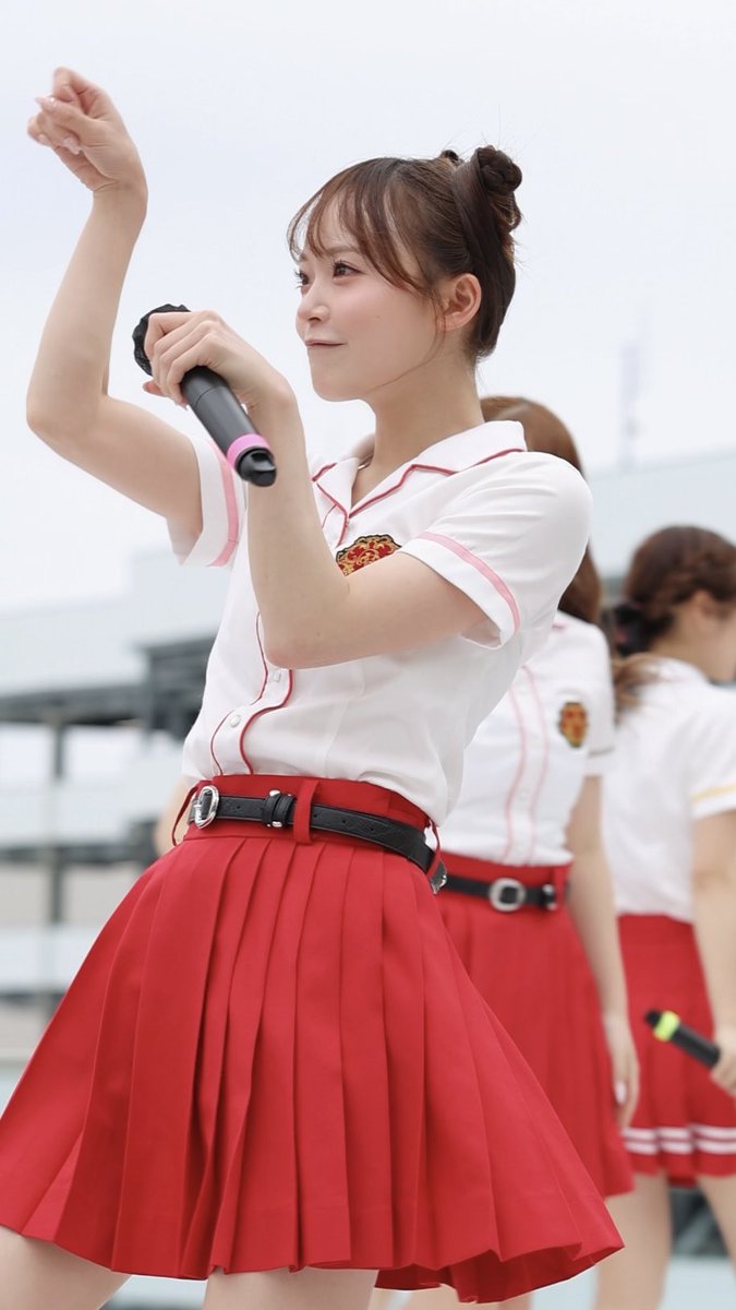 A performer with a high bun hairstyle holds a microphone on stage, wearing a white short-sleeved shirt with pink trim and a red pleated skirt with a black belt.