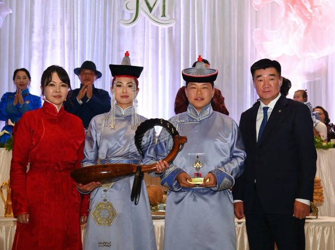 Four people pose at a formal event with 'JM' branding, including a woman in a red traditional dress and two individuals in Mongolian traditional attire presenting awards and a stringed instrument.