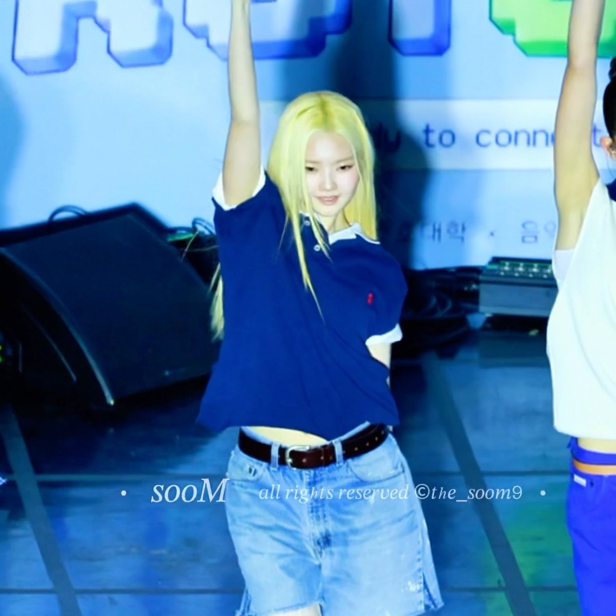 A young woman with blonde hair in a navy polo shirt and denim skirt dancing on stage with her arm raised; watermark reads 'sooM all rights reserved ©the_soom9'.